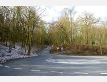 Eine asphaltierte Landstraße führt durch einen winterlichen Wald mit kahlen Bäumen und leicht mit Schnee bedecktem Boden. Mehrere Verkehrsschilder sind am Straßenrand sichtbar, darunter Park- und Warnschilder.