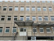Mehrstöckiges Verwaltungs- oder Bürogebäude mit grauer Betonfassade und regelmäßig angeordneten rechteckigen Fenstern. Im Vordergrund befindet sich ein Eingang mit Treppe, weißer Doppeltür und einem gebogenen Metall-Vordach.
