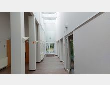 Heller Innenflur eines modernen Gebäudes mit weißen Wänden, Holztüren und großen Fenstern oben, die Tageslicht einlassen. Am Ende des Flurs befindet sich eine gläserne Eingangstür, die nach draußen führt, und seitlich sind teilweise Bürotüren sowie ein kleiner bepflanzter Bereich sichtbar.