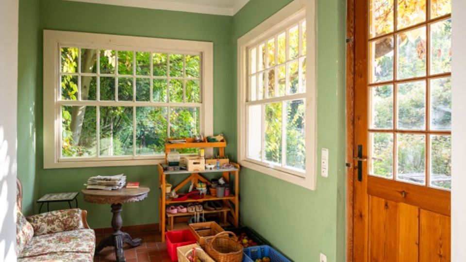 Heller Eingangs- oder Wintergartenraum mit grün gestrichenen Wänden, großen Sprossenfenstern und einer Holztür mit Glaseinsatz. Terrakottafarbener Fliesenboden, kleines Sofa und Tisch sowie ein Regal und Kisten für Gartenutensilien vor Blick ins grüne Außenumfeld.