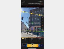 Kreuzung in einer städtischen Umgebung mit einer dunklen Eckfassade eines mehrstöckigen Gebäudes. Im Vordergrund sind Fußgänger, Ampeln und Straßenmarkierungen sichtbar; im Bild auch Sonnenverlauf und Zeitinformationen durch eine App eingeblendet.
