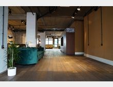 Ein modernes Loft-Interieur mit freiliegender Betondecke, Ziegelwänden und einer Einrichtung aus Holzböden. Im Raum stehen ein grünes Sofa, Pflanzen, eine Wand mit weißer Kachelung sowie große Fenster, die Tageslicht hereinlassen.