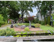 Ein gepflegter Garten mit einer Terrasse aus roten Ziegelsteinen, möbliert mit Gartenmöbeln und einem Kamin. Im Hintergrund stehen ein rotes Backsteinhaus und moderne Wohngebäude, umgeben von großen Bäumen, Hecken und Rasenflächen. Im Vordergrund ist ein Steg an einem Gewässer zu sehen.