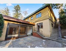 Innenhof eines zweigeschossigen Wohnhauses mit gelber Holzfassade und hellem Ziegel-Sockel, großer Glas-Schiebetürfront und gepflasterter Terrasse. Rechts führen Außentreppen mit Geländer am Hang entlang nach oben, umgeben von Bäumen und Sträuchern.