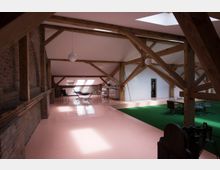 Loftartiger Innenraum mit sichtbaren Holzbalken, Ziegelwänden und vielen Dachschrägen. Der Boden ist teils rosa glänzend, teils mit grünem Teppich bedeckt, und es gibt eine Mischung aus modernen Möbeln, darunter Stühle, ein Tisch und eine Hängematte.