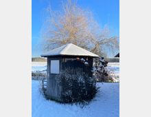 Kleiner hölzerner Pavillon mit schneebedecktem Dach, umgeben von schneebedeckten Sträuchern und karger winterlicher Landschaft. Im Hintergrund steht ein kahler Baum vor einem klaren blauen Himmel.