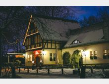 Nächtliche Außenansicht eines historischen Gasthauses im Fachwerkstil mit schneebedecktem Satteldach, beleuchteten Fensterrahmen und einem Eingangsschild „Altes Zollhaus“. Vor dem Gebäude liegt ein verschneiter Hof mit Bänken, Kettenpfosten und kahlen Bäumen im Hintergrund.