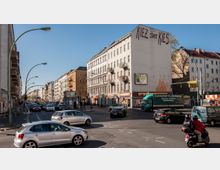 Straßenszene in einem urbanen Wohnviertel mit mehrstöckigen Wohnhäusern aus der Gründerzeit, Geschäften im Erdgeschoss und einem Graffiti-Schriftzug „Kiez statt Kies“ auf einer Fassade. Mehrspurige Straße mit Autos, einem Lkw, einem Roller und Fußgängern bei sonnigem Wetter.