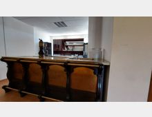 Barbereich in einem schlichten Raum mit hellem Anstrich und Deckenplatten. Der Tresen aus Holz hat eine dunkle, verzierte Front und eine Metalloberfläche, dahinter befinden sich Regale mit Gläsern und Geschirr.