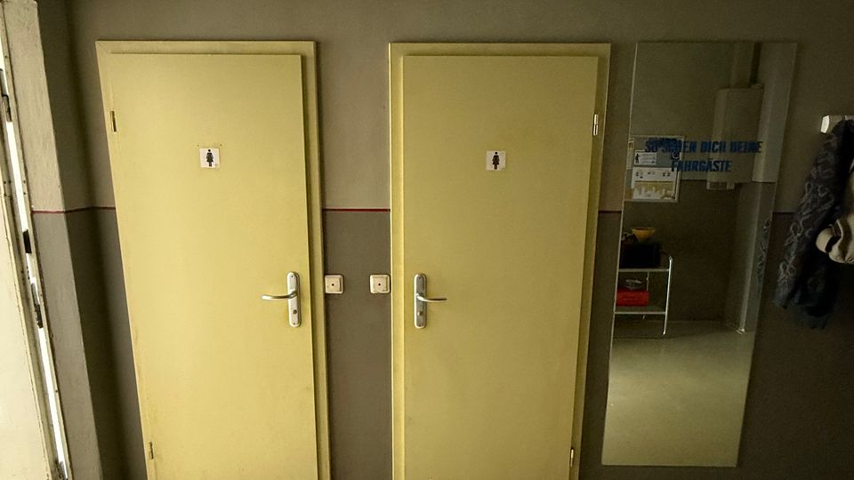 Innenraum eines Flurs mit zwei nebeneinanderliegenden, hellgelben Toilettentüren mit Piktogrammen an einer grauen Wand. Rechts hängt ein großer Wandspiegel neben einer Garderobe; der Boden ist hell und glatt.