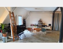 Ein geräumiger, moderner Dachraum mit sichtbaren Holzbalken, einem Glastisch mit mehreren bunten Stühlen, einem hohen Regal voller Utensilien im Hintergrund und einer Holzleiter an der linken Wand. Links steht eine Topfpflanze, und der Boden besteht aus glattem, grauem Beton.