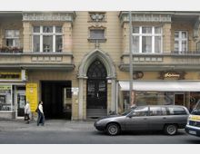 Fassadenansicht eines historischen Wohn- und Geschäftshauses mit gelb-beiger Fassade und verzierten Fenstern. Im Erdgeschoss befinden sich eine KFZ-Werkstatt mit gelbem Schild, ein Durchgang zum Hinterhof, ein Eingang mit dekorativem Rundbogen sowie eine Bäckerei mit weißer Markise. Vor dem Gebäude parken Autos.