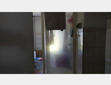 Schmaler Badezimmerflur mit gefliester Wand, der zu einer Dusche mit milchiger Glastür führt. Links ist ein WC-Bereich mit Heizkörper zu sehen, an der Duschwand hängt eine Badematte.