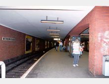 U-Bahn-Station mit einem gebogenen Bahnsteig, umgeben von roten Ziegelwänden. An der Wand ist das Stationsschild "Mehringdamm" angebracht, es gibt beleuchtete Werbetafeln und hängende Leuchten an der Decke.