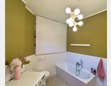 Modernes Badezimmer mit olivgrün gestrichenen Wänden und weiß gefliester Badewanne an der rechten Seite, dazu Waschmaschine und WC links. An der Decke hängt eine kugelförmige Mehrfachleuchte, über dem WC ist ein heruntergezogenes weißes Rollo, der Boden ist hell gefliest.