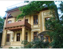 Gelbes, zweistöckiges Wohngebäude aus Backstein mit großen Holzfenstern, einem überdachten Balkon und dekorativen Architekturelementen. Vor dem Haus befinden sich Bäume, Sträucher und ein blühender Rhododendron.