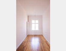 Schmale, helle Innenaufnahme eines leeren Raums mit Holzfußboden, weiß gestrichenen Wänden und einer hohen Decke. Am Ende des Raumes befindet sich ein rechteckiges Fenster mit abgerundeter Oberkante, links daneben ein Heizkörper.