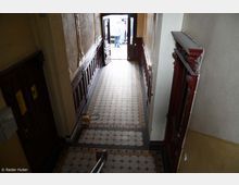 Ein schmaler Altbauflur mit verziertem Fliesenboden und dunklen Holzvertäfelungen an den Wänden. Am Ende des Flurs führen geöffnete Doppeltüren nach draußen, wo Tageslicht und Menschen erkennbar sind.