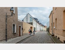 Eine gepflasterte Gasse mit Reihenhäusern in hellen und sandfarbenen Tönen, flankiert von einfachen Fassaden mit Fenstern und Türen. Drei Personen gehen auf dem Gehweg entlang, während im Hintergrund ein moderneres Gebäude mit Giebeldach sichtbar ist.