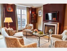 Eleganter Wohnbereich mit cremefarbenen Polstermöbeln, einem Glastisch und einem dekorativen Kamin mit Holzvertäfelung. Im Hintergrund befindet sich ein großes Fenster mit Blick auf das Brandenburger Tor.