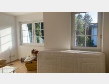 Wohnzimmer mit hellem Laminatboden, zwei Fenstern mit weißen Rahmen und Blick auf einen Garten. Im Vordergrund steht eine Matratze an der Wand, daneben ein gelbes Ledersofa mit Stofftieren, und rechts eine Stehleuchte.