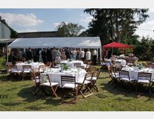 Gartenbereich mit runden, weiß gedeckten Tischen und Holzklappstühlen auf einer Rasenfläche, daneben ein großes weißes Zelt und ein rotes Pavillonzelt. Im Hintergrund stehen hohe Bäume, Büsche und eine niedrige Mauer bzw. Gebäudefassade.