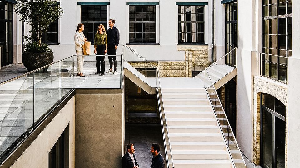 Innenhof eines modernen Bürogebäudes mit hellen Fassaden, großen Sprossenfenstern und dekorativen Ziegelbögen über den Fensterreihen. Eine breite, weiße Treppe und Glasgeländer verbinden zwei Ebenen; unten liegt ein gepflasterter Hofbereich mit Sitzbank und Pflanzkübel.