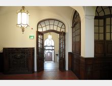 Innenansicht eines historischen Gebäudes mit Holztäfelung und gefliesten Böden. Ein hölzerner Eingang mit Glaselementen führt zu einem Flur mit großen Bogenfenstern, dekorativen Lampen und einer doppelflügeligen Außentür.