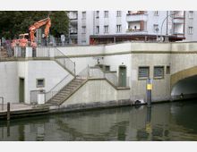 Eine Uferpromenade mit einem Treppenaufgang aus Beton, Geländern und kleinen Fenstern, gelegen an einem Wasserkanal. Im Hintergrund sind mehrstöckige Wohngebäude und ein Baukran zu sehen; rechts führt eine Brücke über das Wasser.
