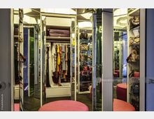 Ein luxuriöser begehbarer Kleiderschrank mit verspiegelten Türen, Regalen mit Designerhandtaschen, hängender Kleidung und einer zentralen, runden, pinkfarbenen Sitzbank. Der Raum ist gut beleuchtet und verfügt über eine edle Inneneinrichtung.