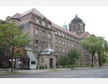 Ansicht eines historischen Verwaltungsg Gebäudes mit einer sandsteinfarbenen Fassade, zahlreichen Fenstern und einem roten Ziegeldach. Im Hintergrund ist eine große Kuppel sichtbar, während im Vordergrund Bäume, ein Zaun und eine leere Straße zu sehen sind.