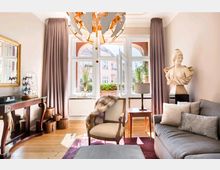 Wohnzimmer mit eleganter Einrichtung: Ein Zimmer mit großen Fenstern, die viel Tageslicht hereinlassen, lila Vorhängen, einer grauen Couch mit Kissen und einem Beistelltisch mit Lampe. Eine Büste steht auf einem Podest, und die Deckenleuchte hat ein auffälliges, modernes Design.