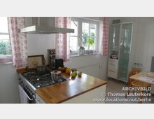 Helle Küche mit großem Gasherd, hölzerner Arbeitsplatte und Dunstabzugshaube, daneben ein Fenster mit rosa geblümten Vorhängen und einer Fensterbank mit Pflanzen und Dekoration. Im Hintergrund ein weißer Vitrinenschrank und ein kleiner Esstisch mit zwei Stühlen.