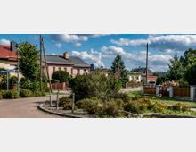 Eine ruhige Wohnstraße in einem Vorort mit Einfamilienhäusern, kleinen Gärten und Gehwegen. Im Vordergrund sind Büsche und Grünflächen zu sehen, während im Hintergrund Strommasten und ein bewölkter blauer Himmel sichtbar sind.