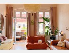 Wohnzimmer mit hellen Wänden, großen Fenstern und Zugang zu einem Balkon mit Holzboden. Im Raum befinden sich eine orangefarbene Sitzbank, ein braunes Ledersofa, ein großer Pflanzenkübel und dekorative Elemente wie ein runder Papierlampenschirm und eine Wandzeichnung.