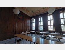 Raum mit Holzverkleidung und hohen rechteckigen Fenstern mit dekorativen Gitterelementen, möbliert mit einfachen Holztischen und Plastikstühlen, unter einer verzierten Decke mit runden, hängenden Leuchten.