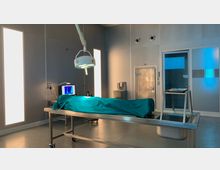 Ein steriler Obduktionsraum mit einem metallenen Seziertisch, auf dem ein Körper unter einem grünen Tuch liegt, einer großen OP-Lampe, einem Computerbildschirm und einer Spüle; im Hintergrund sind eine Tür und beleuchtete Wandelemente zu sehen.