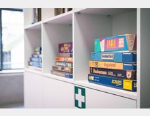 Weiße Regale in einem hellen Innenraum, mit mehreren gestapelten Brettspielen in den Fächern. Unten ist ein Schrank mit einem grünen Erste-Hilfe-Symbol zu sehen, daneben ein großes Fenster im Hintergrund.