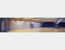 Panoramabild eines modernen Mehrzweckraums mit hellem Holzfußboden und Wänden aus Sichtbeton, teilweise dunkelblau gestrichen. Der Raum ist spärlich ausgestattet mit Stuhlreihen an den Wänden, einer Fluchttür und großen Spiegeln auf der rechten Seite.