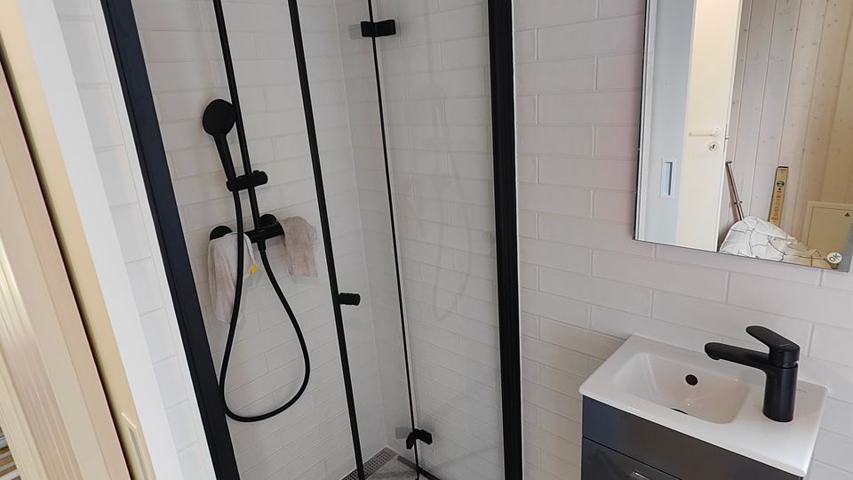 Modernes Badezimmer mit bodengleicher Dusche und Glasabtrennung, weißen Wandfliesen und grauem Fliesenboden. Schwarze Duscharmatur mit Regendusche sowie ein kleiner Waschtisch mit Spiegel und schwarzem Wasserhahn sind zu sehen.