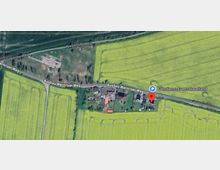 Luftaufnahme eines ländlichen Gebiets mit gelb blühenden Feldern und einem kleinen Weiler entlang der Straße "Ausbau Wernitzer Weg". Einzelne Gebäude mit roten und grauen Dächern sind von Gärten umgeben, und kleinere Wege durchziehen die Landschaft.