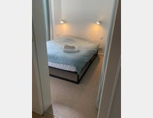 Ein kleines Schlafzimmer mit einem Doppelbett, das eine blaue Bettdecke hat. Zwei Wandleuchten sorgen für Beleuchtung, und auf dem Bett liegen gefaltete Handtücher; der Boden ist mit Teppich ausgelegt.