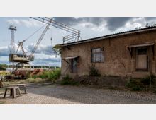 Eine rustikale Industrieumgebung mit einem alten Gebäude aus braunem Putz, das zwei einfache Eingänge und ein Holzfenster hat. Daneben steht ein großer Kran, und im Vordergrund sind Kopfsteinpflaster sowie etwas Vegetation sichtbar.