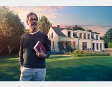 Ein Mann mittleren Alters mit Brille hält ein Buch in der Hand und steht vor einem großen weißen Einfamilienhaus mit mehreren Fenstern und einem gepflegten Garten. Im Hintergrund sind Bäume und ein klarer Himmel zu sehen, während die Sonne tief steht.