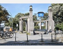 U-Bahn-Eingang Heidelberger Platz in Berlin, mit einem halbrunden Säulenbau aus Stein, einer blauen Hinweistafel mit dem Buchstaben „U“, sowie Laternen auf den Säulen. Im Vordergrund befinden sich ein Gehweg, abgestellte Fahrräder und Straßenelemente; im Hintergrund sind Bäume und parkende Autos zu sehen.
