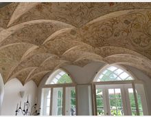 Räumlicher Innenraum mit einem bemalten Kreuzgewölbe, verziert mit floralen Mustern in sanften Farben. Unterhalb des Gewölbes befinden sich große Fenster und eine Glastür mit geschwungenen Sprossen, die den Blick ins Grüne freigeben.