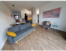 Wohnbereich in einer modernen Wohnung mit Holzboden, einer grauen Couch mit gelben Kissen, einem grauen Sessel mit buntem Kissen, einem abstrakten Wandbild und einer offenen Küche im Hintergrund mit Holzdetails. Am linken Bildrand befindet sich ein Esstisch aus Holz mit Hockern.