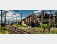 Bahnstrecke mit mehreren Gleisen und Oberleitungen, umgeben von Vegetation und historischen Gebäuden mit Ziegeldächern; im Hintergrund ein Zug und Strommasten. Die Szene spielt sich bei Tageslicht unter einem leicht bewölkten Himmel ab.