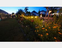 Beleuchteter Garten in der Dämmerung mit breiten Blumenbeeten und einem Rasenweg, gesäumt von Lichterketten und kleinen Bodenleuchten. Im Hintergrund stehen mehrere Wohnhäuser mit Satteldächern sowie Bäume und Sträucher.