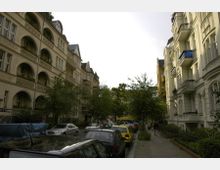 Wohnstraße in einer Stadt mit historischen Mehrfamilienhäusern im Jugendstil auf beiden Seiten. Geparkte Autos säumen die Straße, und vereinzelt sind Bäume entlang des Gehwegs zu sehen.